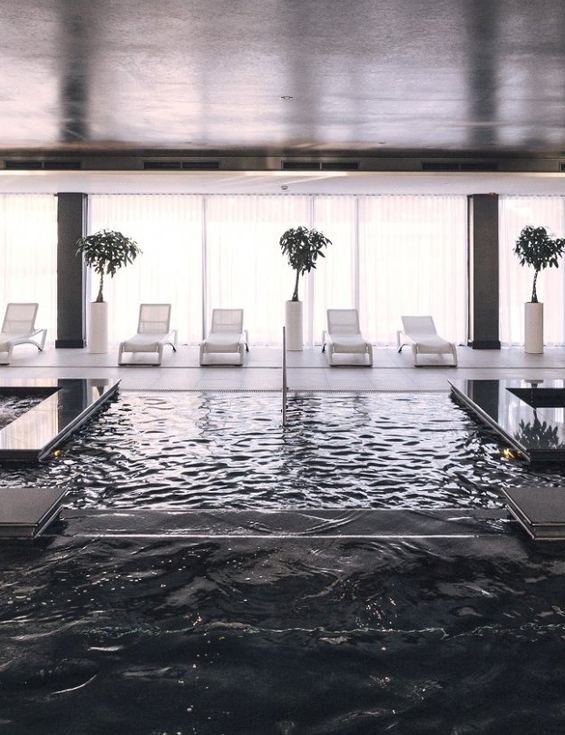 Indoor luxury spa pool with dark reflective water, four large gold pillars and white loungers by floor-to-ceiling windows
