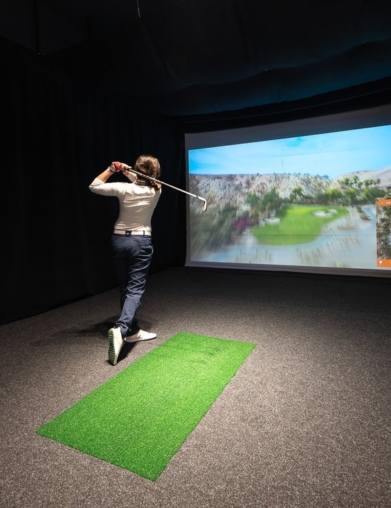Guest swinging a club in a private indoor golf simulator room with projected course, putting mat and Titleist bag.