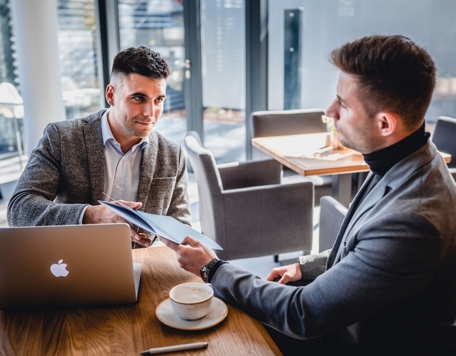 Two men in a hotel cafe having a business meeting, sharing documents over a laptop and coffee at a sunlit table by large windows.