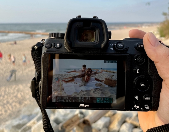 Camera screen showing a family posing on a sandy beach picnic with blanket, wicker basket and sea view.