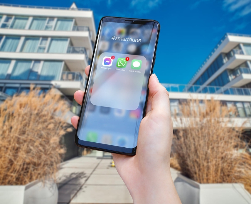 Hand holding smartphone with messaging apps and #smartdune, set before a modern hotel with glass balconies and sunlit terrace.