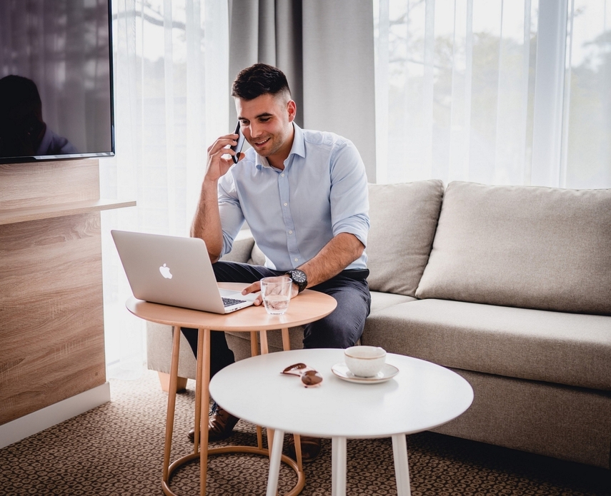 Business traveler on a sofa using laptop and phone in a bright modern hotel suite, round tables with coffee cup and glass of water.