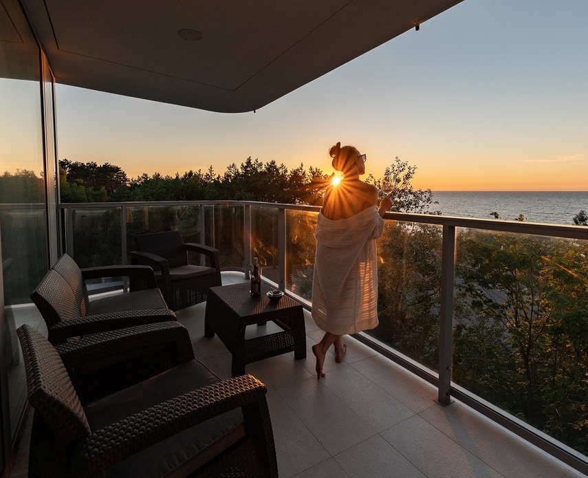 Guest wrapped in a towel on a hotel balcony at sunset, glass railing, sea view, wicker seating and a wine glass