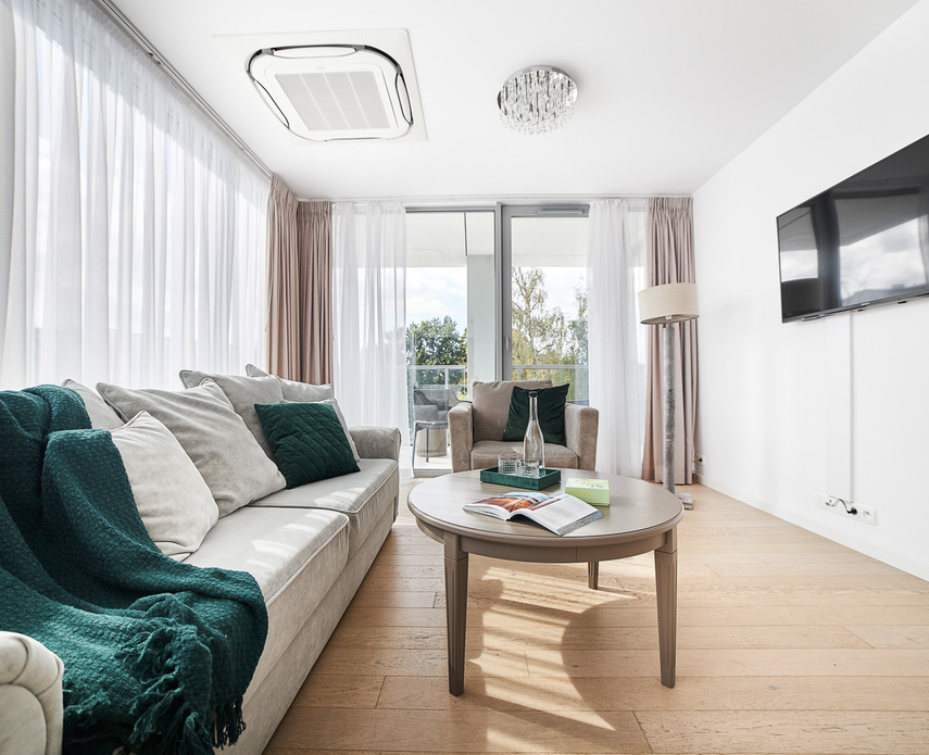 Bright hotel-suite living area with beige sofa, teal throw and cushions, armchair, coffee table, balcony view and wall TV