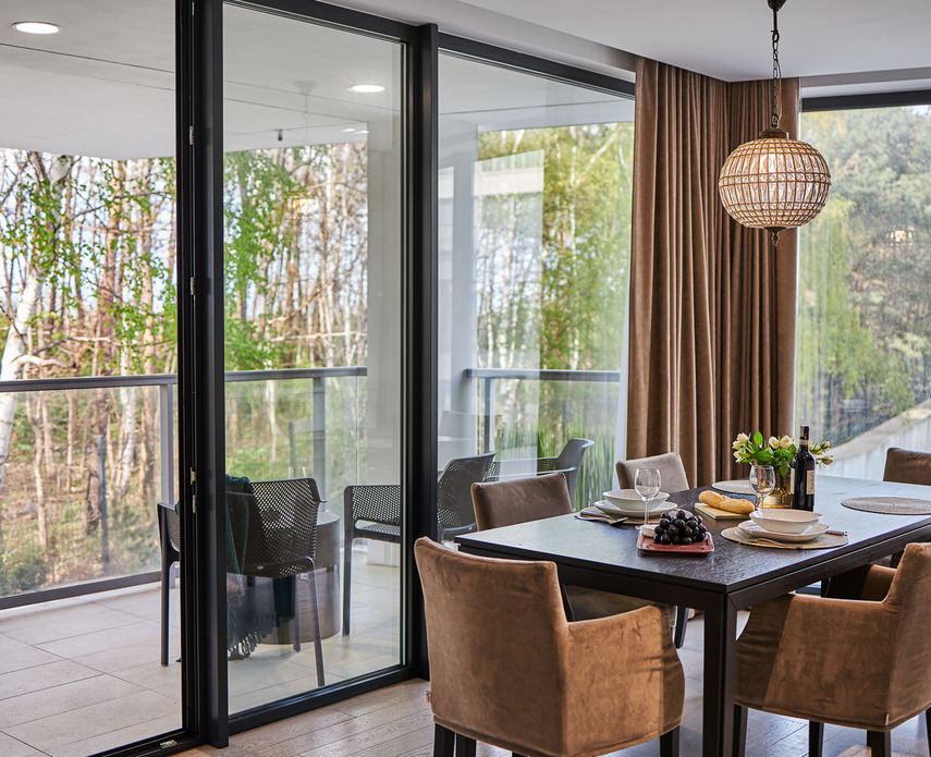 Dining area with dark wood table set for four, velvet chairs, pendant light and balcony with forest view through floor-to-ceiling windows.