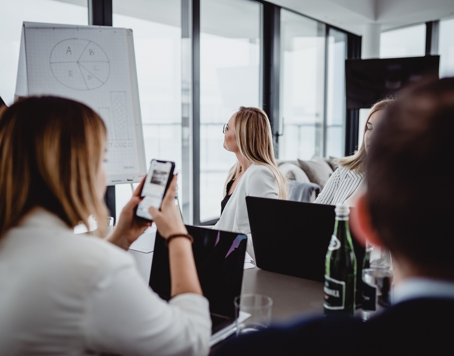 Bright modern meeting room with floor-to-ceiling windows, presenter at flipchart, attendees with laptops and bottled water