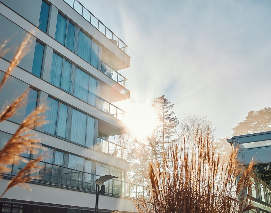 Sunlit modern hotel exterior with glass-railed balconies, floor-to-ceiling windows and decorative grasses by a tranquil terrace.