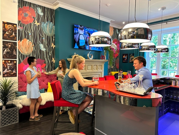 Vibrant hotel lounge with red lacquer bar, chrome pendant lights, floral wallpaper and guests enjoying drinks by a fireplace.