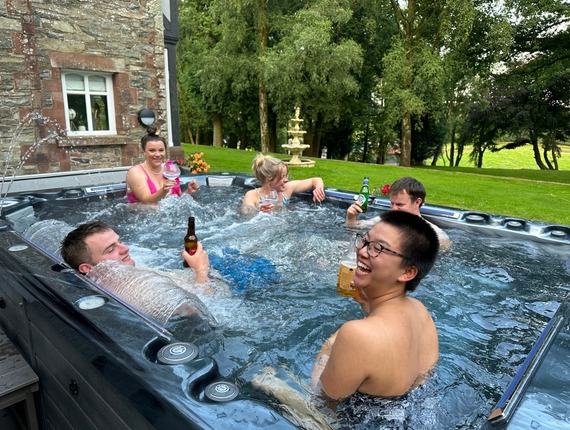 Guests laughing and sipping drinks in an outdoor hot tub beside a stone manor, with a manicured lawn, fountain and trees.