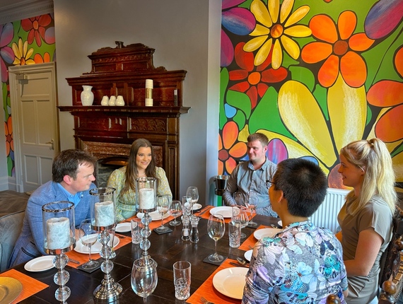 Guests seated around a table in a boutique hotel's dining room with floral mural, antique wood fireplace and crystal candleholders.