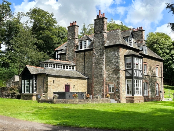 Historic stone country manor with tall chimneys, bay and dormer windows, lawn-front terrace with potted flowers and surrounding mature woodland.