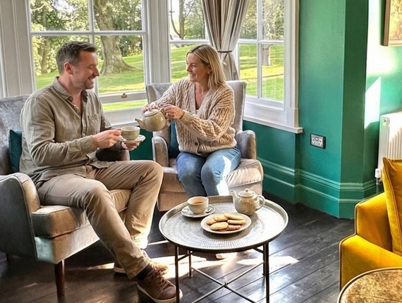 Couple enjoying tea and biscuits in a cozy hotel lounge with emerald walls, velvet armchairs and yellow sofa, garden views.