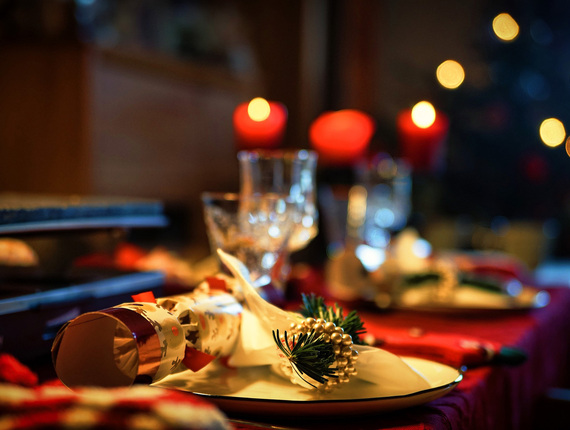 Candlelit holiday table set with crackers, plates, crystal glasses and a napkin ring of evergreen sprig and gold beads, warm bokeh lights.