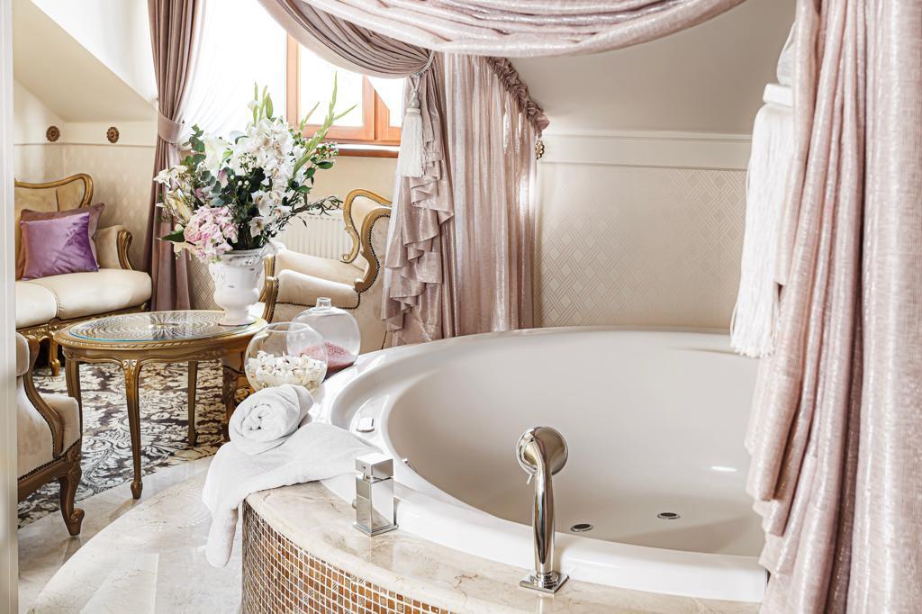 Luxurious circular bathtub beside ornate seating and gold-accent table, vase of flowers, soft pink drapes.