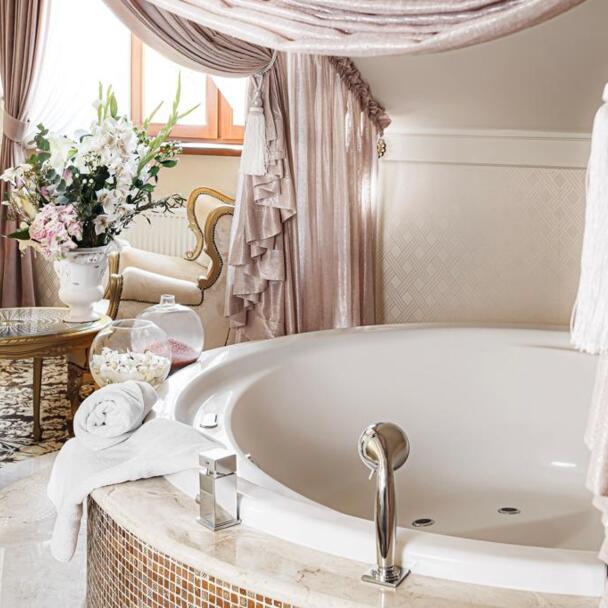 Round jetted bathtub with rolled towel and chrome faucet, pink drapery and floral arrangement, elegant.