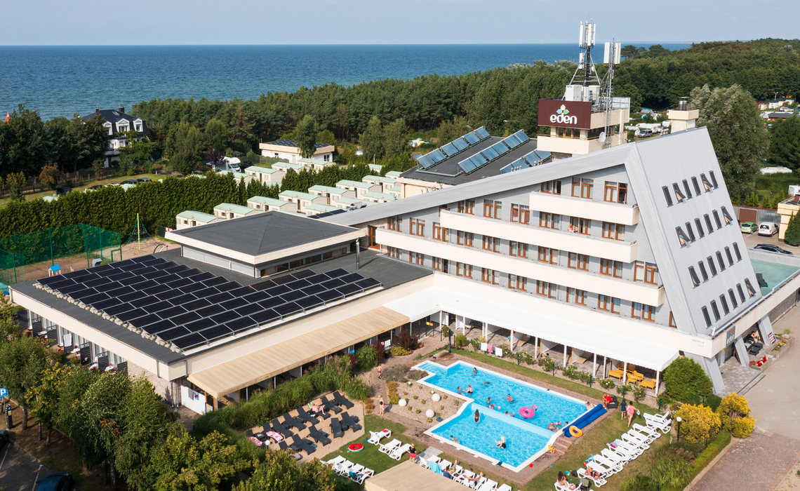 Nadmorski hotel ze skośnym skrzydłem i panelami słonecznymi, z widokiem na morze, odkrytym basenem z kąpiącymi się i leżakującymi gośćmi.