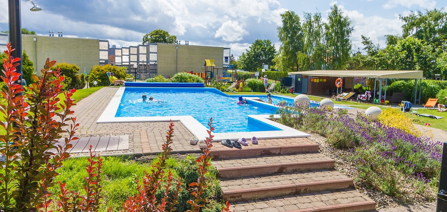 Open hotel pool with a terrace, loungers and a covered relaxation area; children swimming, lavender beds and brick steps.