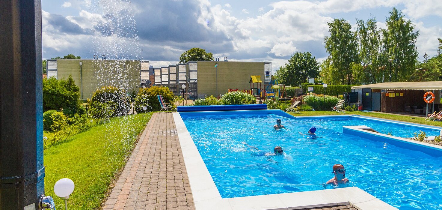Hotel's outdoor pool with light-blue water, children swimming, an outdoor shower, loungers and a playground in the background