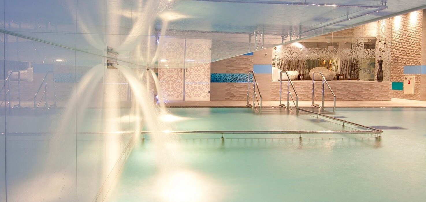 Indoor spa pool with water cascades, steps into the water and sunbeds, modern tiles and cozy lighting