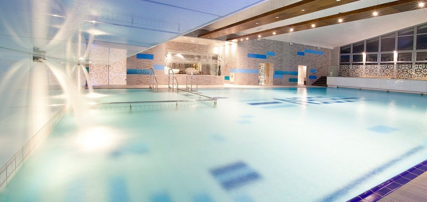 Indoor hotel pool with water cascades, entry steps, modern decor, soft blue lighting and large windows