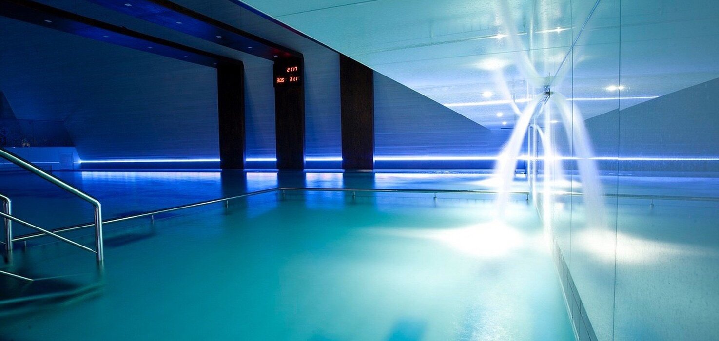 Indoor spa pool with blue mood lighting, water cascades, steps and a metal handrail.