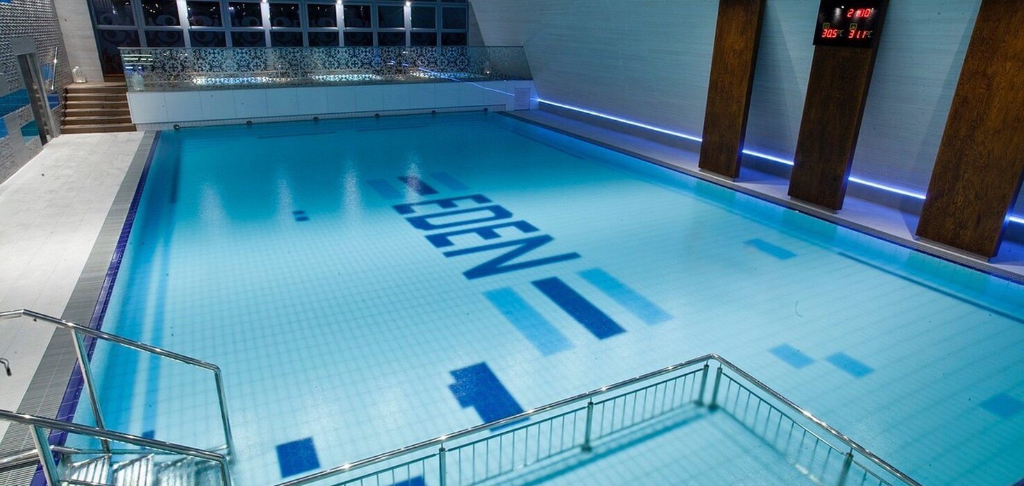 Interior of an indoor hotel pool with illuminated blue water, steel stairs, a decorative partition and the word EDEN on the bottom.