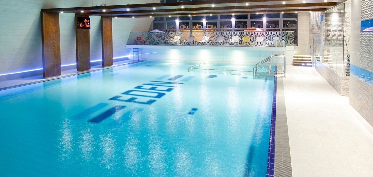 Spacious, softly lit indoor hotel pool with turquoise water, wooden beams, a mosaic balcony and steel steps