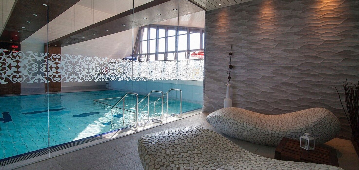Indoor hotel pool behind a glass wall with a decorative band, metal steps, designer loungers and a wavy wall texture