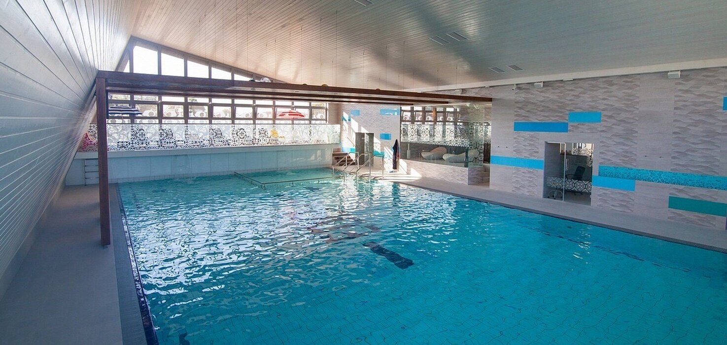 Indoor hotel swimming pool with turquoise tiles, sloped wood-paneled ceiling, sunlit windows, glass partition and pool steps.
