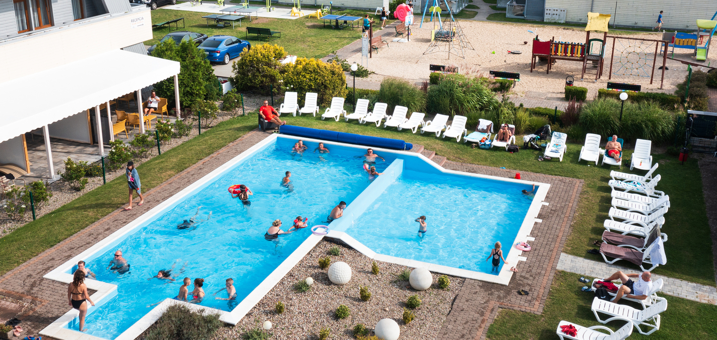 Hotel outdoor pool with families swimming, a row of loungers, a covered relaxation area and a playground in the background