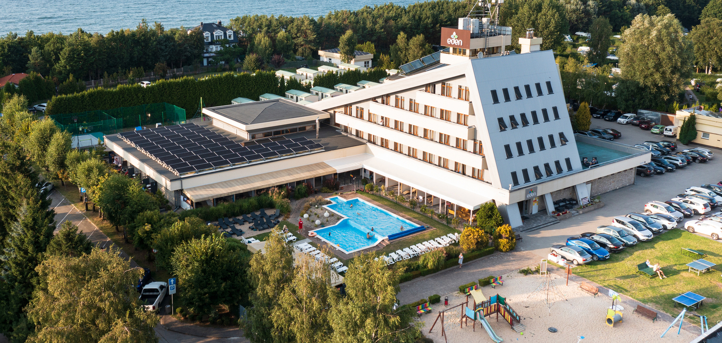 Coastal hotel with angled modern façade, rooftop solar panels, outdoor pool with loungers, sandy kids' playground and sea view.
