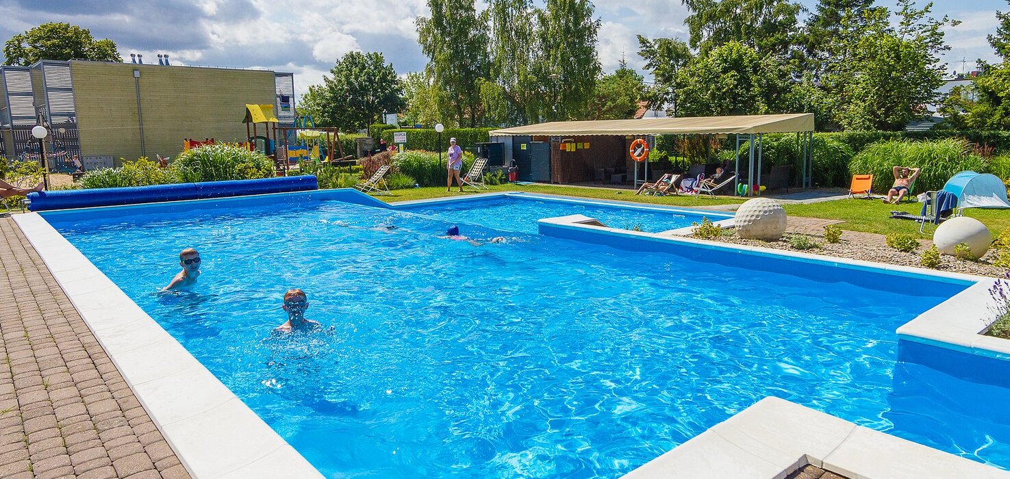 Hotel outdoor pool with crystal-blue water, loungers, a covered patio, a playground and children swimming.