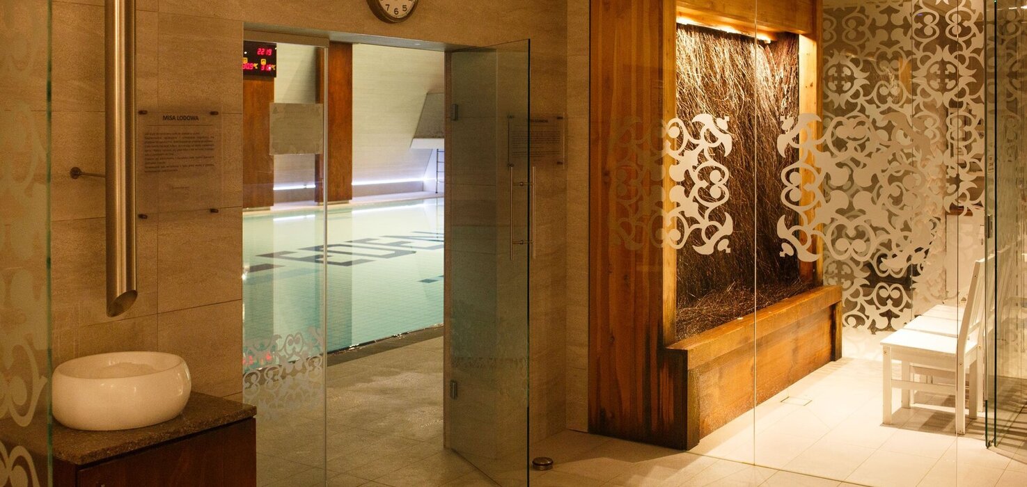 Indoor pool behind glass doors, decorative matte patterns, a wooden water wall, white benches and warm lighting.