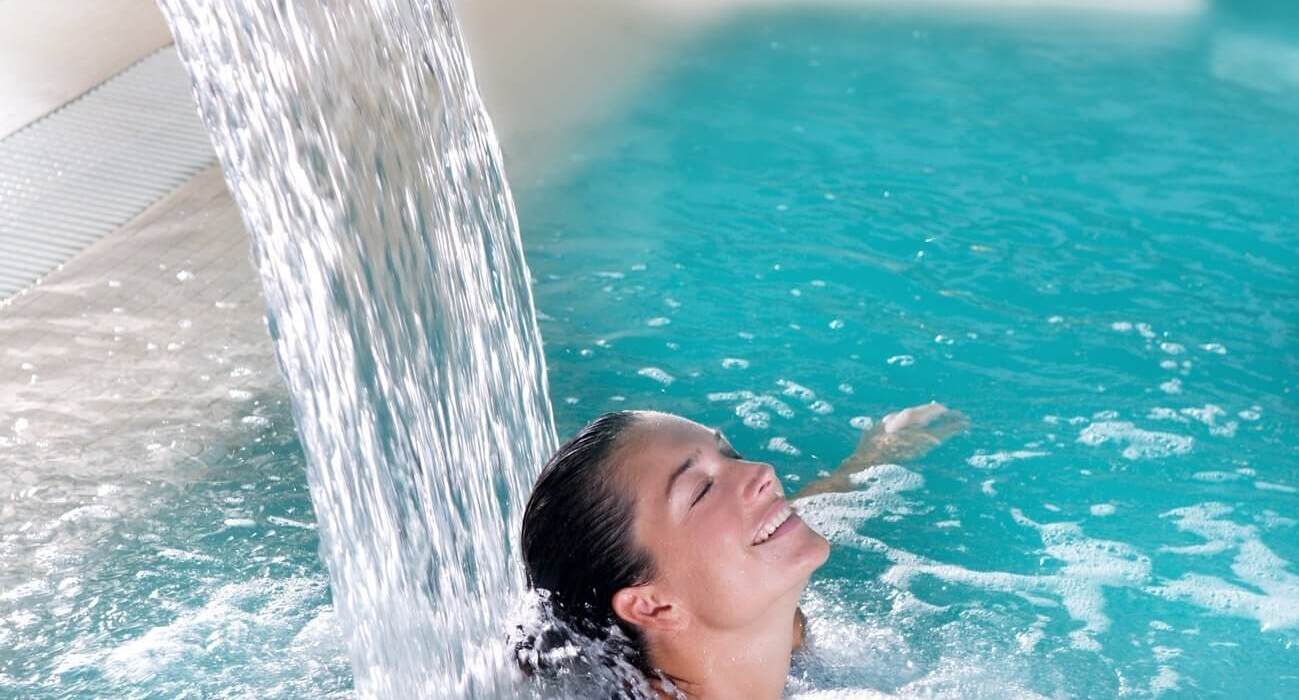 A woman relaxing under a massage water jet in a blue hotel spa pool, bubbles and a waterfall