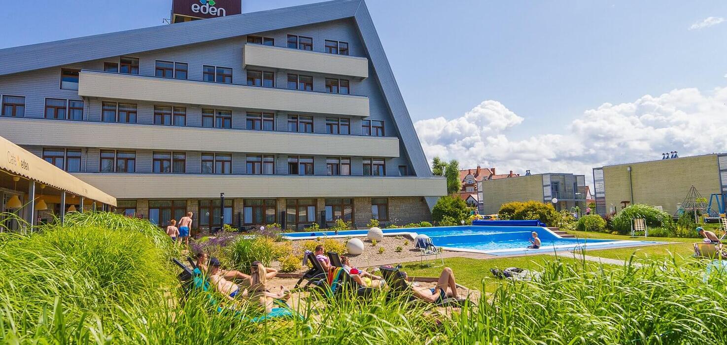 Hotel with a slanted roof and terraced balconies, an outdoor pool, loungers on the lawn and guests sunbathing on a sunny day.