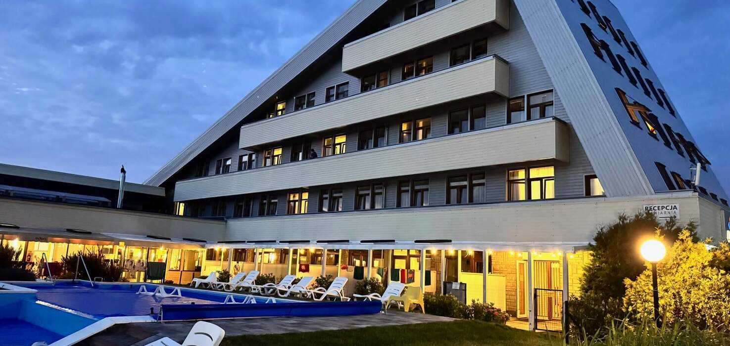 Triangular hotel with terraced floors and lit rooms, an evening pool by the building with loungers and a reception.