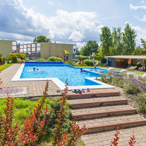 Open hotel pool with a terrace, loungers and a covered relaxation area; children swimming, lavender beds and brick steps.