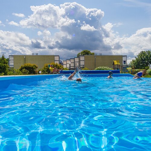 Outdoor hotel pool with crystal-blue water, guests swimming, loungers and a playground by the buildings