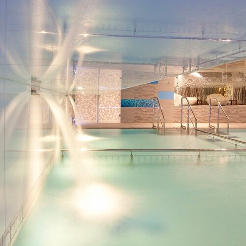 Indoor spa pool with water cascades, steps into the water and sunbeds, modern tiles and cozy lighting