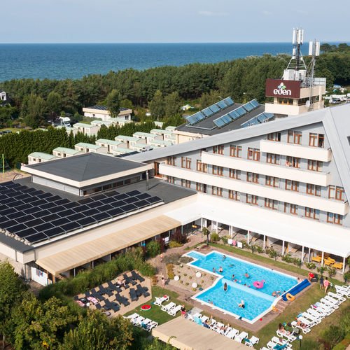 Seaside hotel with a slanted wing and solar panels, sea view, an outdoor pool with guests swimming and sunbathing.