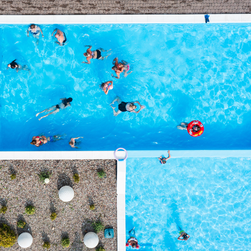 Hotel complex of two pools with light-blue water, families swimming, stone paving with a flower bed