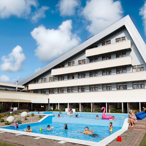 Hotel with a slanted roof and terraced balconies, outdoor pools with families, an inflatable flamingo and loungers.