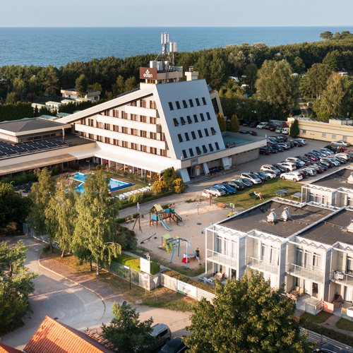 Seaside hotel with a distinctive slanted wing, an outdoor pool, a playground, balconies and parking, with views of the sea and forest.