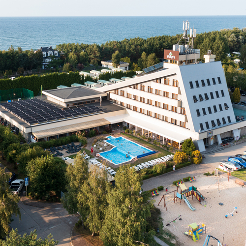 Coastal hotel with angled modern façade, rooftop solar panels, outdoor pool with loungers, sandy kids' playground and sea view.