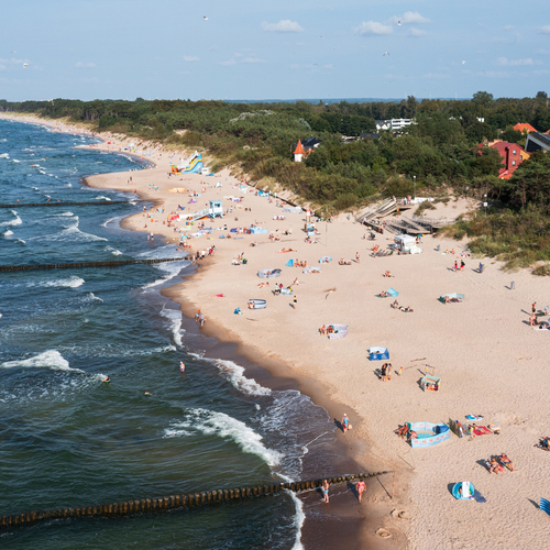 Sandy beach with breakwaters, guests swimming and sunbathing, dunes, a coastal forest and hotels in the background.