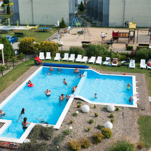 Open hotel pool with loungers, guests swimming and sunbathing, a small garden and a playground in the background