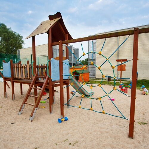 A sandy playground with a wooden tower, a slide, a rope climbing net, a swing and toys; a child playing nearby.