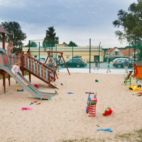 A family fenced sandy playground by the hotel with a wooden slide, spring riders and a sports field in the background.