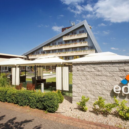 Hotel Eden with a slanted roof, balconies with plants, shaded relaxation pavilions, a well-kept garden and a logo.
