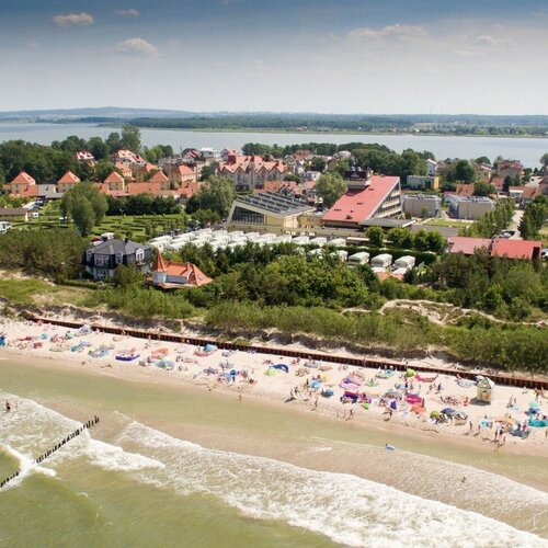 Beach with a sandy shore and colorful umbrellas, a waterfront hotel complex with red roofs, dunes, wooden groynes and a bay in the background
